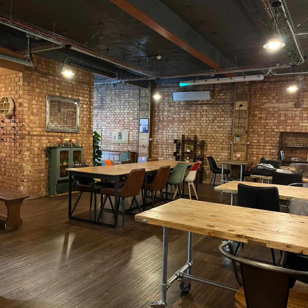Several long wood-topped tables surrounded by chairs, on a brown floor and with a brick wall, shelves and a mirror in the background, in the first floor at Understated coffee shop.Â